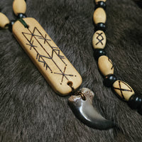 A close-up of a beaded necklace laid on dark fur. The necklace has light natural wood beads with black engraved symbols, black round beads, a large rectangular wooden pendant with engraved geometric designs, and a dark gray curved bone or horn charm.