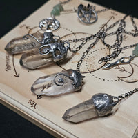 Several clear crystal point pendant necklaces with silver-toned chains and caps are arranged on a light wood display board against a dark background.