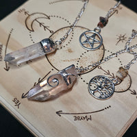 Three silver-toned crystal and metal pendant necklaces displayed on a wooden board with engraved astrological symbols. The necklaces include a clear pointed crystal, a rose-gold-toned crystal pendant, a silver pentagram, and a silver tree of life charm.