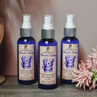 Three bottles of Monster Spray! Monster Spray! essential oil spray, each with a blue label featuring a cartoon monster. The bottles are placed on a wooden surface with a pink flower next to them.