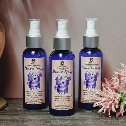 Three bottles of Monster Spray! Monster Spray! essential oil spray, each with a blue label featuring a cartoon monster. The bottles are placed on a wooden surface with a pink flower next to them.